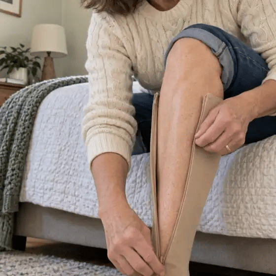 A woman sitting on her bed zips a beige compression sock up her leg